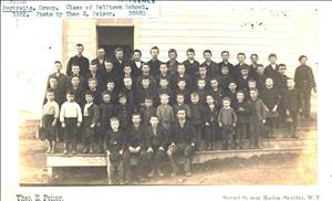 A large group of young students stand on a porch in front of a white, clapboard building, which is the Belltown School. They stand in five rows, with the tallest in the rear and the shortest in front
